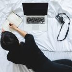 person-writing-in-notebook-beside-laptop-working-from-bed